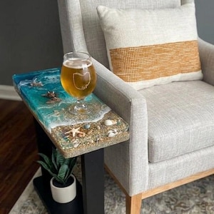 May include: A rectangular side table with a black metal frame and a resin tabletop featuring a beach scene with blue ocean waves, white foam, sand, and seashells. A clear glass with a golden beverage sits on the surface, and a small green plant is on the lower shelf.