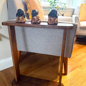 May include: A wooden side table with a dark brown finish, featuring a shelf and a gray fabric panel. Three small black and gold Buddha statues sit on top. The table is placed in front of a gray armchair.