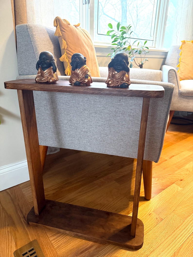 May include: A wooden side table with a dark brown finish, featuring a shelf and a gray fabric panel. Three small black and gold Buddha statues sit on top. The table is placed in front of a gray armchair.