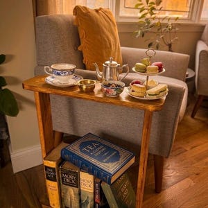 May include: A wooden side table with a tea set, pastries, and books. The tea set includes a silver teapot, a teacup, and small bowls. A two-tiered plate holds macarons and sandwiches. Books include "Tea: A Global History" and "Moby Dick."