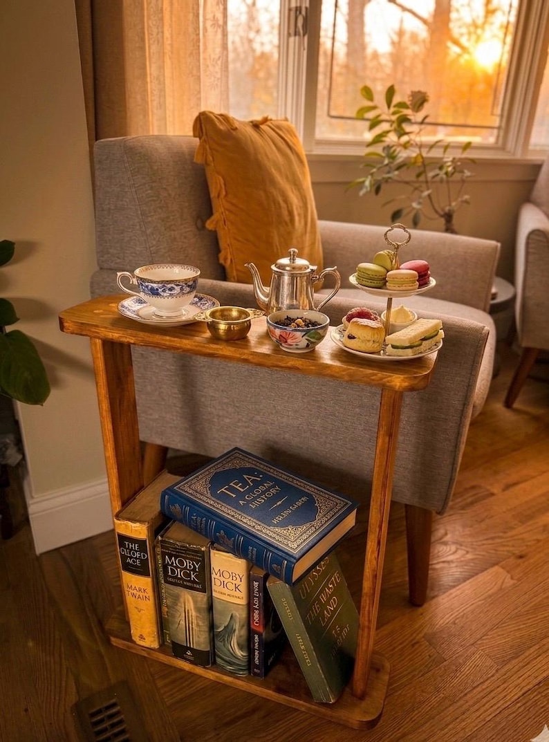 May include: A wooden side table with a tea set, pastries, and books. The tea set includes a silver teapot, a teacup, and small bowls. A two-tiered plate holds macarons and sandwiches. Books include "Tea: A Global History" and "Moby Dick."