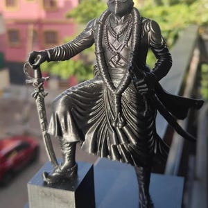 May include: A black monochromatic figurine of a warrior standing on a square base. The figure wears traditional attire with a draped lower garment, a necklace, and a turban. One hand rests on the hilt of a sword, while the other holds a scabbard.