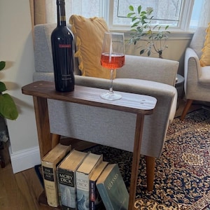 May include: A dark wooden side table with a bottle of red wine and a glass of rosé. The table has a lower shelf with several books. The background includes a window, a gray armchair, and a patterned rug.
