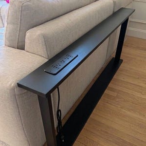 May include: A long, narrow black console table positioned behind a sofa. The table features a flat rectangular surface with an integrated power strip containing multiple electrical outlets and USB ports, supported by a sturdy black metal frame base.