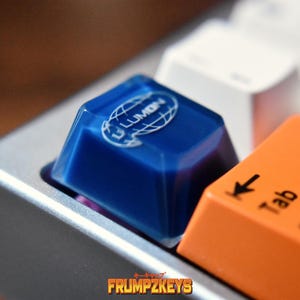 May include: Close-up of a mechanical keyboard with custom keycaps. A blue keycap with the text "LUMON" and a globe design is next to an orange "Tab" key. The keyboard has a silver frame. The logo "FRUMPKEYS" is at the bottom.