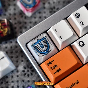Könnte beinhalten: Nahaufnahme einer Computertastatur mit individuellen Tastenkappen. Die Tastenkappen weisen verschiedene Designs auf, darunter ein blau-weißes Emblem, ein Porsche-Logo und ein Guitar Hero-Design. Orange und weiße Tasten sind ebenfalls sichtbar.