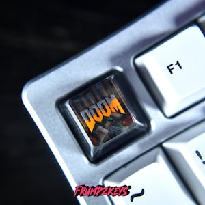 May include: Close-up of a computer keyboard with a custom keycap featuring the word "DOOM" in orange and a graphic. The keycap is surrounded by white keys and a silver frame. The brand name "FRUMPZKEYS" is visible.