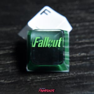 Può includere: Un copritasto personalizzato con la scritta "Fallout" in verde. Il copritasto ha una forma quadrata con una parte superiore verde trasparente e un copritasto "F" bianco dietro. Per tastiere meccaniche.