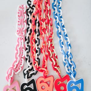May include: A collection of colorful chain necklaces with matching number pendants. The necklaces are in shades of pink, black, red, orange, and blue. Each pendant displays the number "67" in a corresponding color. The chains are made of large, interlocking links.