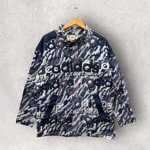 May include: A vintage Adidas sweatshirt with a blue and white abstract print featuring the Adidas logo repeated throughout. The sweatshirt has a mock neck and a kangaroo pocket.