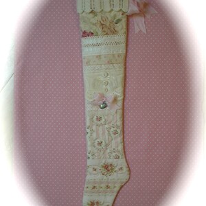 May include: A vintage-style Christmas stocking with a patchwork design. It features floral patterns, lace, and ribbons in cream, pink, and beige. A small silver bell and pink bow are attached, adding a touch of charm. The stocking is approximately 50 cm in length.