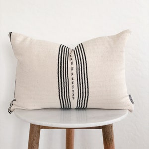 May include: Cream-colored rectangular pillow with a central vertical stripe design in black. The stripe is composed of parallel lines and decorative stitching. The pillow is placed on a small, round, white marble-topped table with wooden legs.
