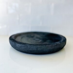 May include: A black, round, wooden dish with a slightly raised rim. The dish is made of dark wood and has a smooth, polished finish.