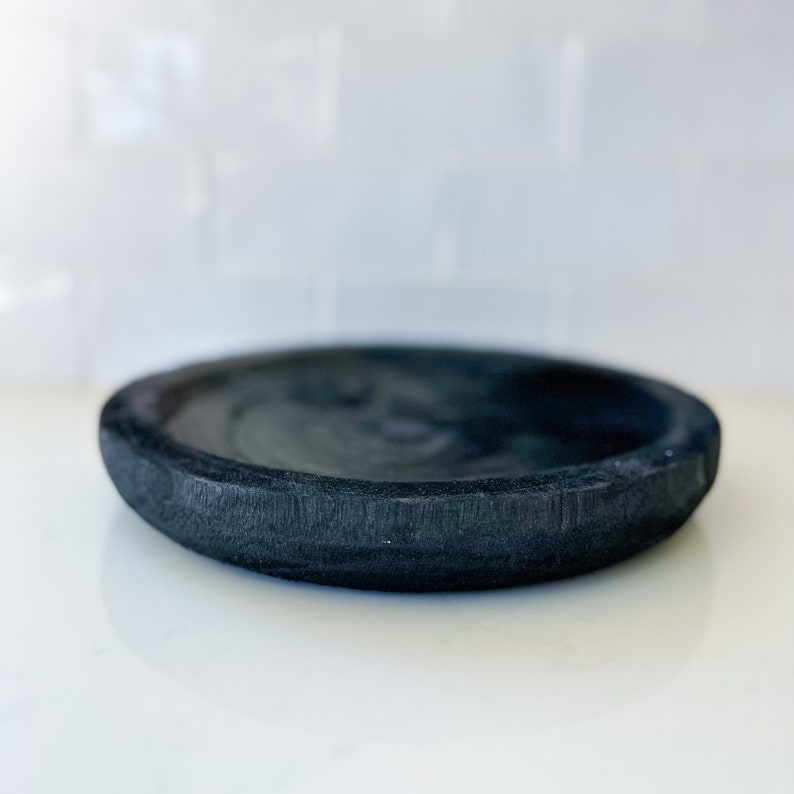 May include: A black, round, wooden dish with a slightly raised rim. The dish is made of dark wood and has a smooth, polished finish.