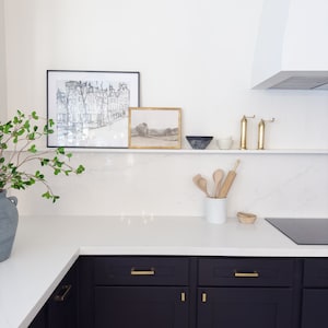 Pu&ograve; includere: Un piano di lavoro da cucina bianco con armadietti neri e ferramenta dorata. Sopra il piano di lavoro, uno scaffale bianco con due disegni in bianco e nero incorniciati, un disegno di paesaggio incorniciato, un mattarello di legno e cucchiai di legno in un barattolo di ceramica bianco. C'&egrave; un piano cottura nero e una ciotola di ceramica bianca sullo scaffale.
