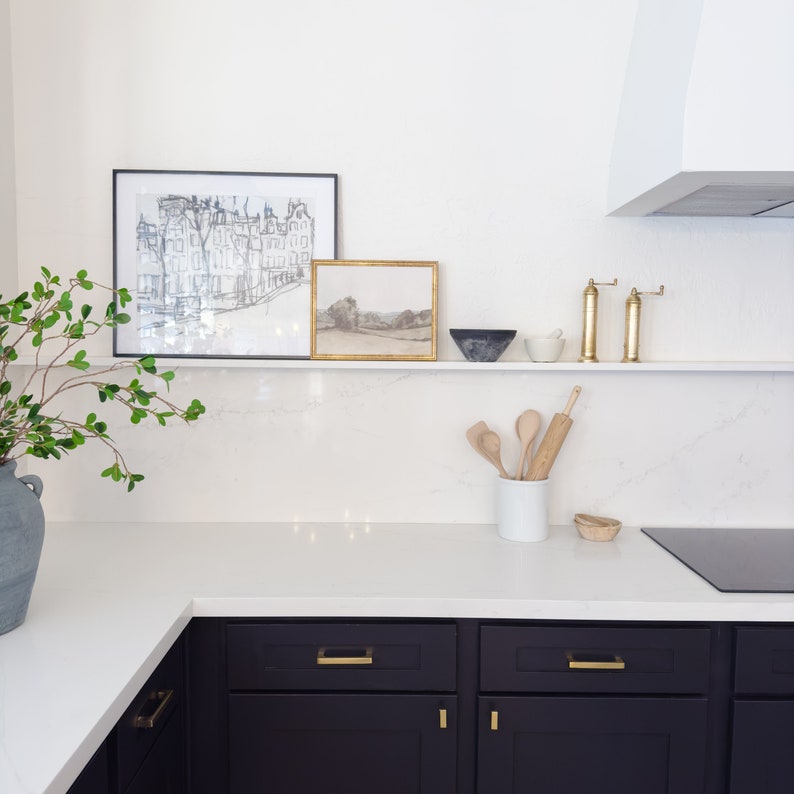 Pu&ograve; includere: Un piano di lavoro da cucina bianco con armadietti neri e ferramenta dorata. Sopra il piano di lavoro, uno scaffale bianco con due disegni in bianco e nero incorniciati, un disegno di paesaggio incorniciato, un mattarello di legno e cucchiai di legno in un barattolo di ceramica bianco. C'&egrave; un piano cottura nero e una ciotola di ceramica bianca sullo scaffale.