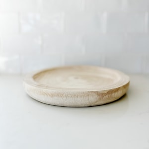 May include: A round wooden tray with a light brown color and a slightly raised edge. The tray is sitting on a white surface.