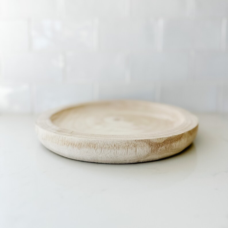 May include: A round wooden tray with a light brown color and a slightly raised edge. The tray is sitting on a white surface.