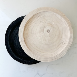 May include: Two round wooden trays, one black and one natural wood. The natural wood tray has a circular grain pattern.