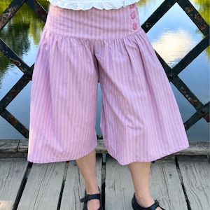 May include: Wide-leg culottes in a light pink and white vertical pinstripe pattern. The garment features a high, fitted waistband with three small pink buttons on the right side and a gathered, voluminous silhouette that falls below the knee.