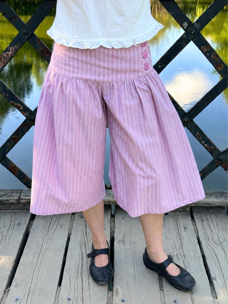 May include: Wide-leg culottes in a light pink and white vertical pinstripe pattern. The garment features a high, fitted waistband with three small pink buttons on the right side and a gathered, voluminous silhouette that falls below the knee.