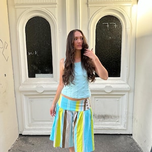 May include: A light blue sleeveless top with vertical seam detailing paired with a knee-length skirt featuring a wide waistband and vertical stripes in shades of bright lime green, light blue, white, and brown, finished with black Mary Jane shoes.