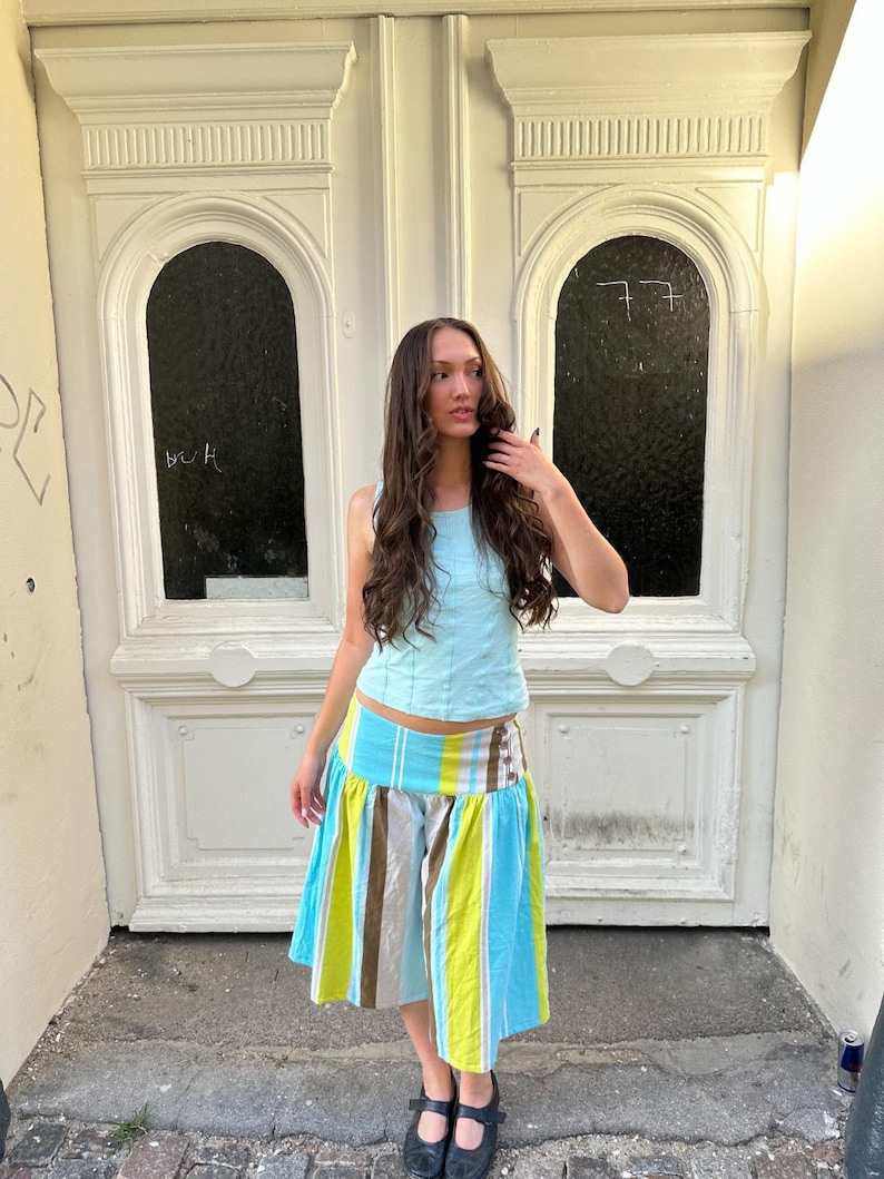 May include: A light blue sleeveless top with vertical seam detailing paired with a knee-length skirt featuring a wide waistband and vertical stripes in shades of bright lime green, light blue, white, and brown, finished with black Mary Jane shoes.