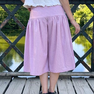 May include: A pair of light pink and white striped culottes featuring a high-waisted design with a gathered, voluminous skirt-like silhouette that falls to mid-calf length. The garment is paired with a simple white top and black shoes.