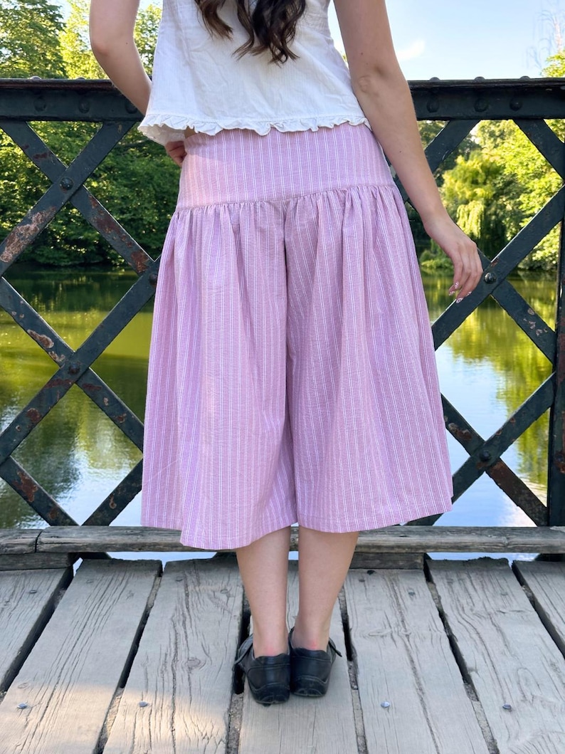 May include: A pair of light pink and white striped culottes featuring a high-waisted design with a gathered, voluminous skirt-like silhouette that falls to mid-calf length. The garment is paired with a simple white top and black shoes.
