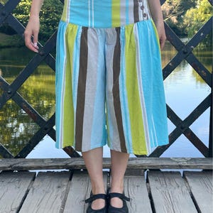 May include: Wide-leg culottes featuring a vertical striped pattern in shades of bright blue, lime green, brown, and white. The garment has a high-waisted band with three dark buttons on the side and a gathered, voluminous silhouette that falls below the knee.