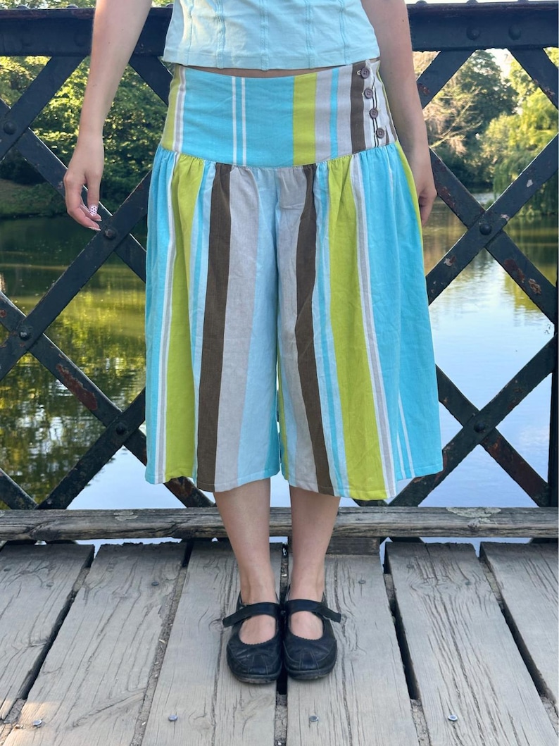 May include: Wide-leg culottes featuring a vertical striped pattern in shades of bright blue, lime green, brown, and white. The garment has a high-waisted band with three dark buttons on the side and a gathered, voluminous silhouette that falls below the knee.