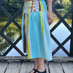May include: A midi-length skirt featuring vertical stripes in shades of light blue, lime green, brown, and white. The skirt has a wide, fitted waistband with a side button closure and a flared, pleated lower section that falls below the knees.