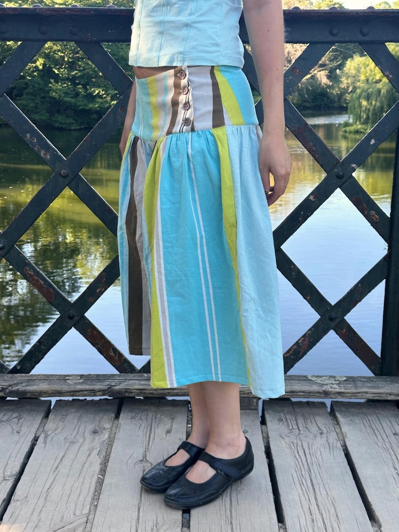 May include: A midi-length skirt featuring vertical stripes in shades of light blue, lime green, brown, and white. The skirt has a wide, fitted waistband with a side button closure and a flared, pleated lower section that falls below the knees.