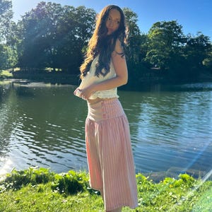 May include: A white sleeveless top with a ruffled hem paired with a high-waisted, calf-length skirt featuring vertical pink and white stripes, a wide waistband, and two visible buttons on the side, worn with black flat shoes.
