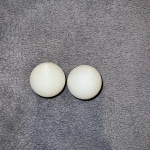 May include: Two white golf balls with dimpled surfaces are positioned closely together. The balls are set against a textured, gray background, creating a simple, yet focused composition. The golf balls are a standard size.