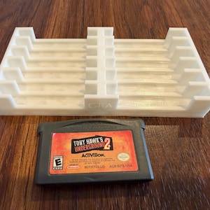 May include: A white 3D-printed game cartridge holder with multiple slots, and a black game cartridge labeled "Tony Hawk's Underground 2" with a red and orange label. The holder is on a wooden surface.