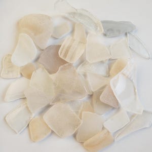 May include: A collection of sea glass pieces in various shapes and sizes. The glass fragments are primarily in shades of white, cream, and pale peach, with some pieces showing subtle textures and patterns. The pieces are scattered, creating a natural, organic aesthetic.