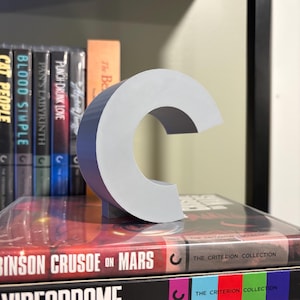 May include: A gray 3D printed letter C, sitting on top of two Criterion Collection DVDs. The DVDs are titled "Robinson Crusoe on Mars" and "Videodrome".
