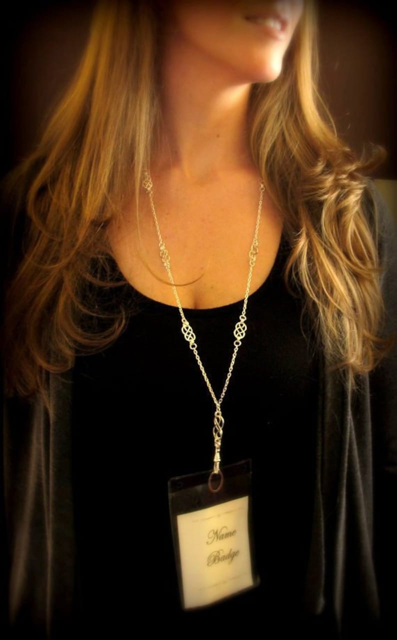 May include: A silver chain necklace with a decorative knot design and a clear plastic badge holder with the text "Name Badge".