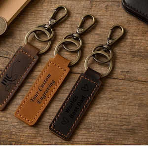 May include: Three rectangular leather keychains with bronze-toned metal lobster clasps and rings. The left keychain features the letters mc, the centre reads Your Custom Engraving, and the right reads Worlds Best Dad with heart icons.