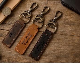 May include: Three rectangular leather keychains with bronze-toned metal lobster clasps and rings. The left keychain features the letters mc, the center reads Your Custom Engraving, and the right reads Worlds Best Dad with heart icons.