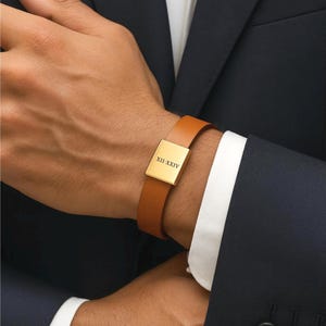 May include: A tan leather bracelet featuring a gold-coloured rectangular plate. The plate is engraved with the Roman numerals XII-XXIV. The bracelet is worn on a wrist, with a dark blue suit jacket and white shirt cuff in the background.