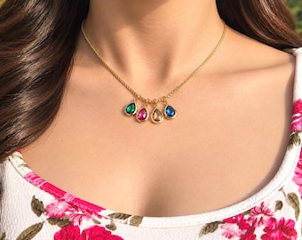 May include: A gold-toned chain necklace featuring four teardrop-shaped pendants in green, pink, champagne, and blue. The necklace rests against a white fabric garment with a pink and green floral pattern, showcasing a delicate metal link design.
