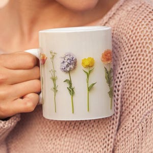 May include: White ceramic mug featuring a floral design with various colorful flowers. The mug has a handle and is held by a person wearing a light pink sweater. The flowers include purple, yellow, and orange blooms.