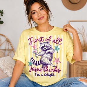 May include: A pale yellow short-sleeve t-shirt featuring a graphic of a raccoon holding a daisy, surrounded by colorful stars and vertical stripes. The text reads, First of all Jesus thinks I'm a delight, in purple script and block lettering.