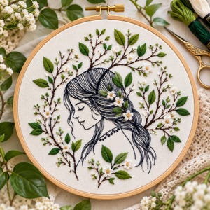May include: A circular wooden embroidery hoop containing white fabric with a black thread outline of a person in profile, surrounded by embroidered green leaves and small white flowers. The design features intricate hair and floral branches.
