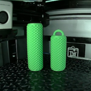 May include: Two green, textured cylindrical objects with a diamond pattern. One is taller, the other shorter with a handle. They are on a black surface, possibly 3D printed. The background includes a 3D printer.