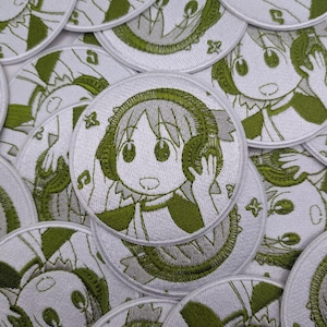 Op de afbeelding: Een verzameling wit en groen geborduurde patches. Elke ronde patch toont een stripfiguur met een koptelefoon, met muzieknoten en bloemmotieven. Het ontwerp is gedetailleerd met groen garen op een witte achtergrond.