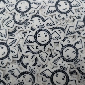 Puede incluir: Una colección de parches de ángeles de fieltro blanco con detalles bordados en negro. Cada parche presenta una cara sonriente, alas y un halo. Los parches están dispersos sobre una superficie blanca.