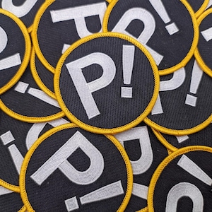 May include: Round black patches with a yellow trim, each displaying a white "P!" and an exclamation mark. The patches are closely arranged, showing a detailed surface. The design is simple and bold, with a clear font.